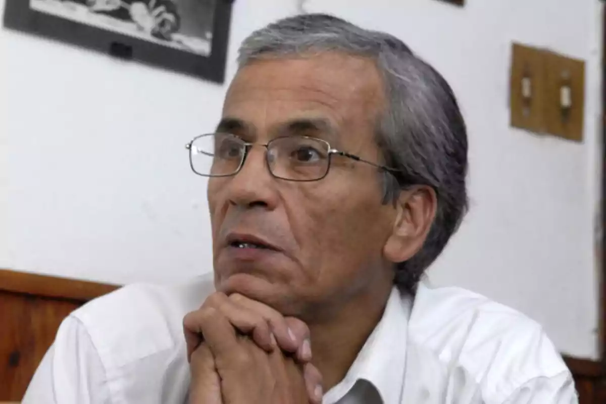 Gray-haired man with glasses, hands clasped under his chin, looking to the side in an indoor room