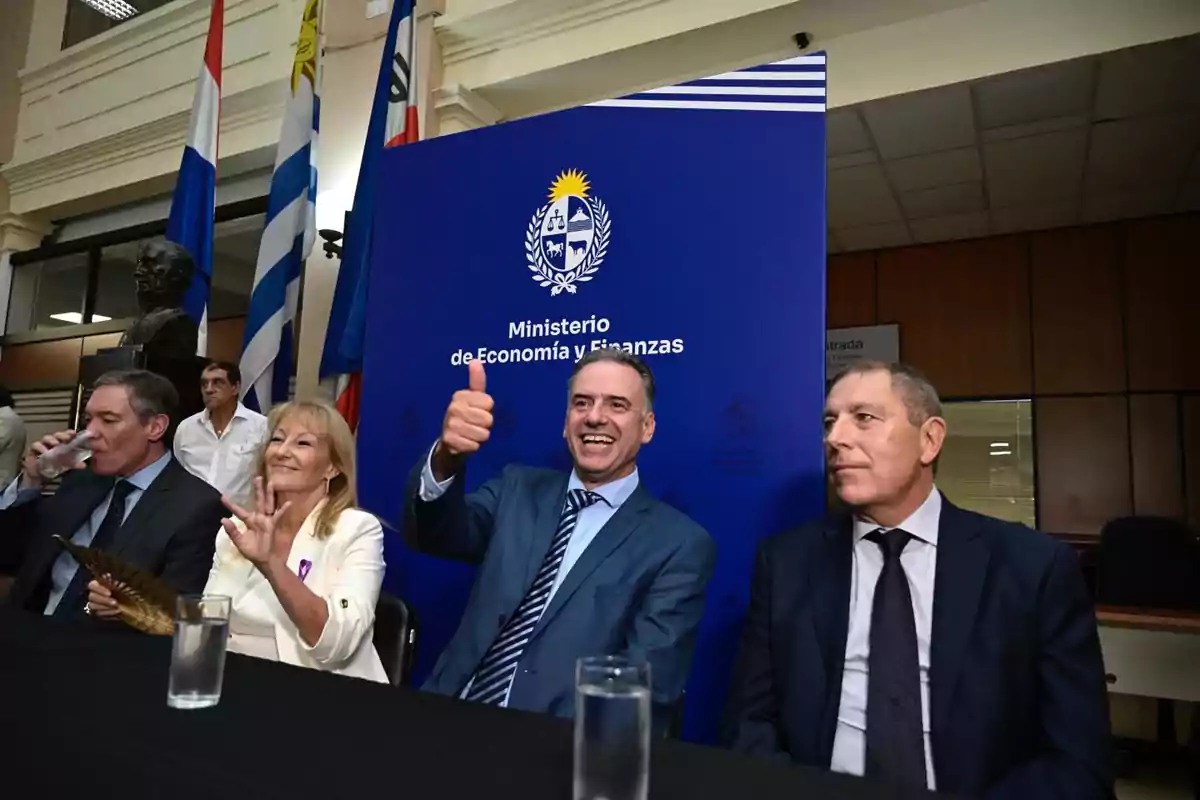Cuatro personas sentadas en una mesa frente a un cartel azul del Ministerio de Economía y Finanzas, una de ellas sonríe y levanta el pulgar en señal de aprobación, mientras que al fondo se ven banderas y una escultura.