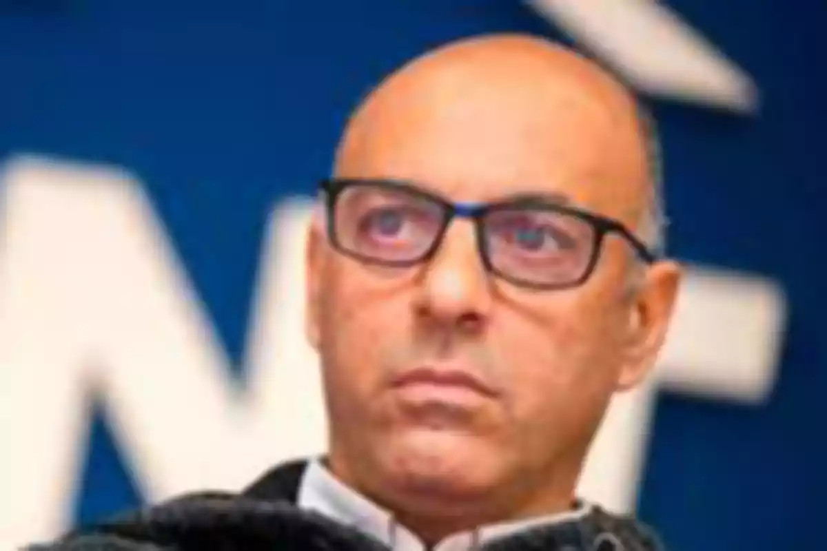 Hombre calvo con gafas y expresión seria frente a un fondo azul desenfocado Hombre calvo con gafas y expresión seria frente a un fondo azul desenfocado