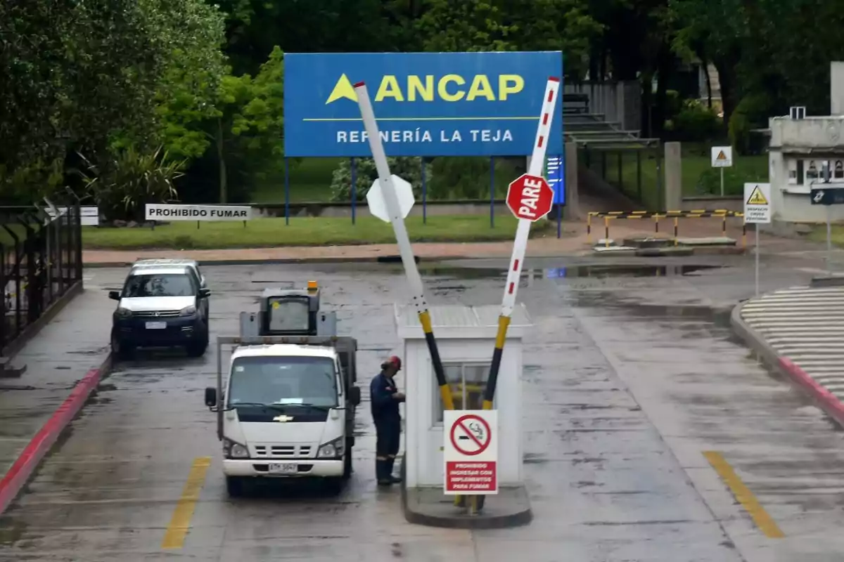 Entrance of the La Teja refinery of ANCAP with a raised barrier, a stopped truck, and a worker next to a control booth, surrounded by traffic signs and warnings