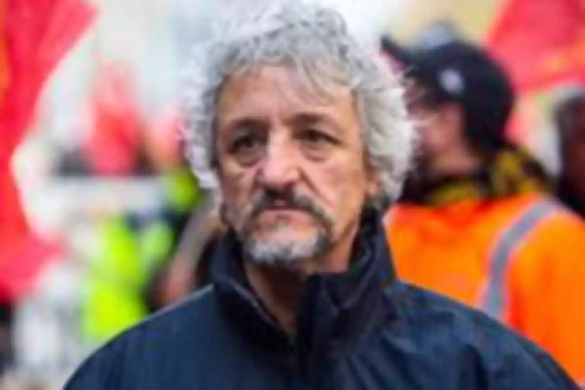A gray-haired man with a beard in an urban setting with people and flags in the background.