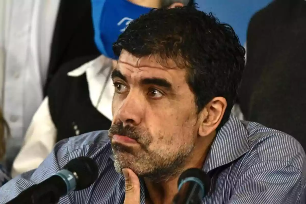 Dark-haired, bearded man with a thoughtful expression sitting in front of two microphones at a press conference Dark-haired, bearded man with a thoughtful expression sitting in front of two microphones at a press conference