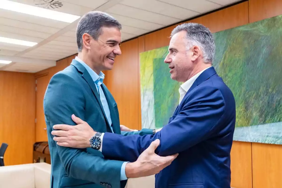 Dos hombres de traje se saludan cordialmente en una oficina con paredes de madera y un cuadro abstracto al fondo.