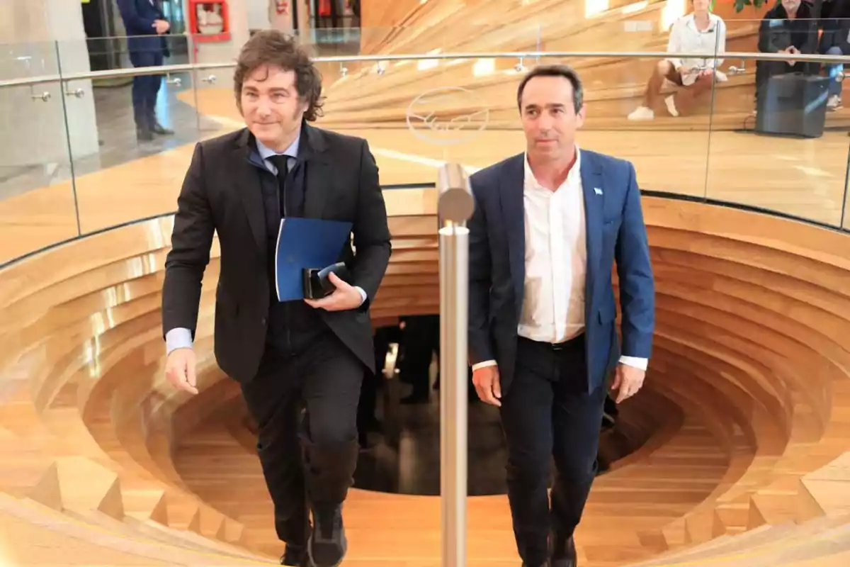 Dos hombres con traje suben una escalera de madera en un edificio moderno con barandales de vidrio y personas sentadas al fondo. Dos hombres con traje suben una escalera de madera en un edificio moderno con barandales de vidrio y personas sentadas al fondo.