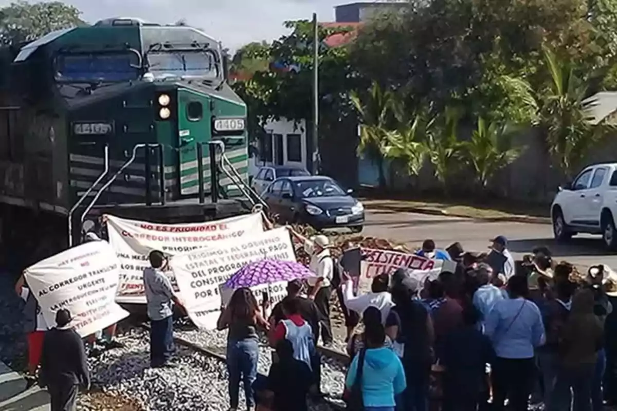 A group of people blocks the train tracks with signs and blankets while a locomotive is stopped in front of them in an urban area surrounded by trees and cars. A group of people blocks the train tracks with signs and blankets while a locomotive is stopped in front of them in an urban area surrounded by trees and cars.