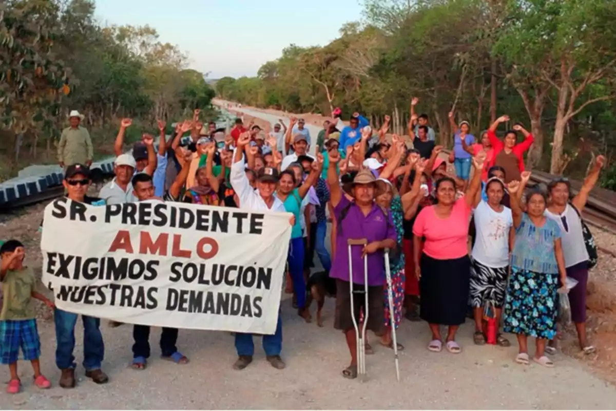 A large group of people is gathered next to some train tracks, raising their fists and holding a banner that calls on President AMLO to address their demands, surrounded by vegetation. A large group of people is gathered next to some train tracks, raising their fists and holding a banner that calls on President AMLO to address their demands, surrounded by vegetation.