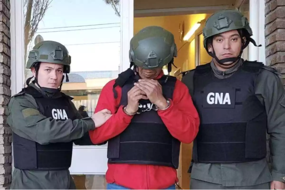 Tres personas con chalecos antibalas y cascos, dos de ellas con uniforme de la GNA, escoltan a una persona con sudadera roja y esposas.