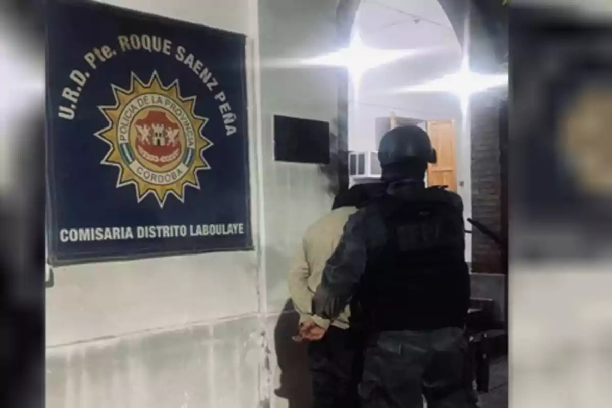 Un oficial de policía escolta a una persona esposada hacia el interior de una comisaría, junto a un cartel que indica "Comisaría Distrito Laboulaye" de la Policía de la Provincia de Córdoba.
