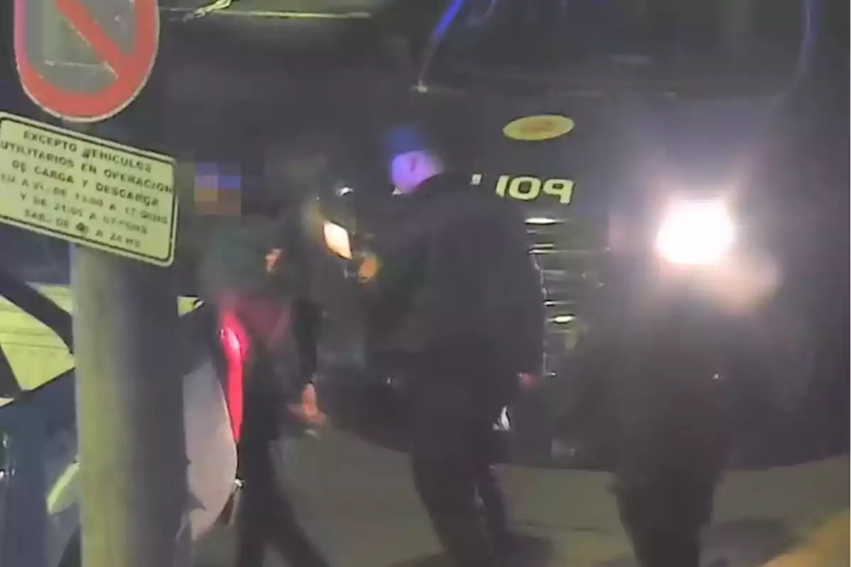 Policías interactúan con una persona junto a una patrulla en una calle de noche, con un letrero de tránsito visible en primer plano.