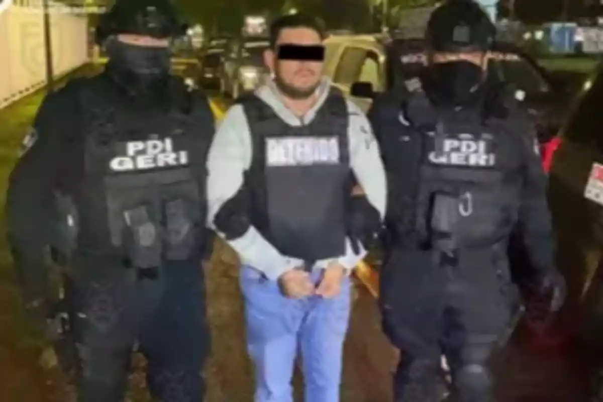 A handcuffed man wearing a "detainee" vest is escorted by two police officers in tactical gear.