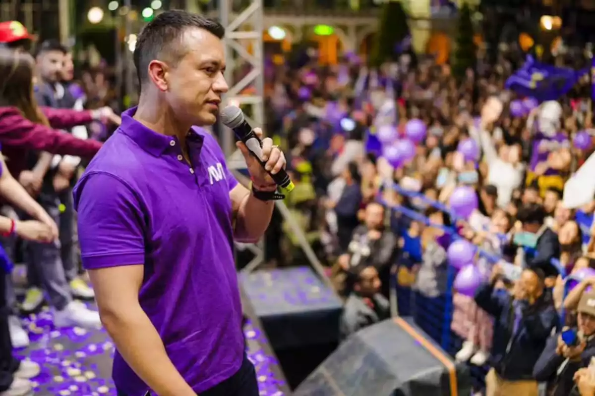 A man in a purple jersey speaks into a microphone in front of a crowd at an outdoor event.