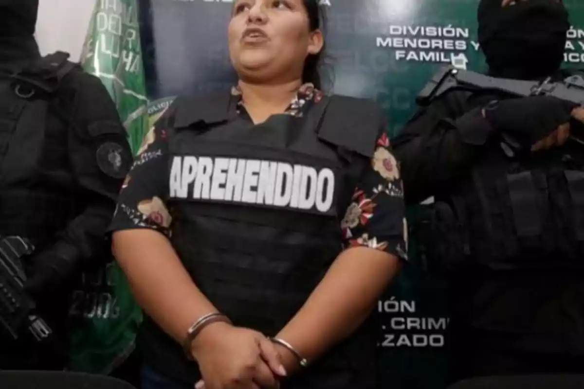 Una persona con chaleco que dice "APREHENDIDO" está esposada y flanqueada por dos agentes de seguridad en un entorno oficial.