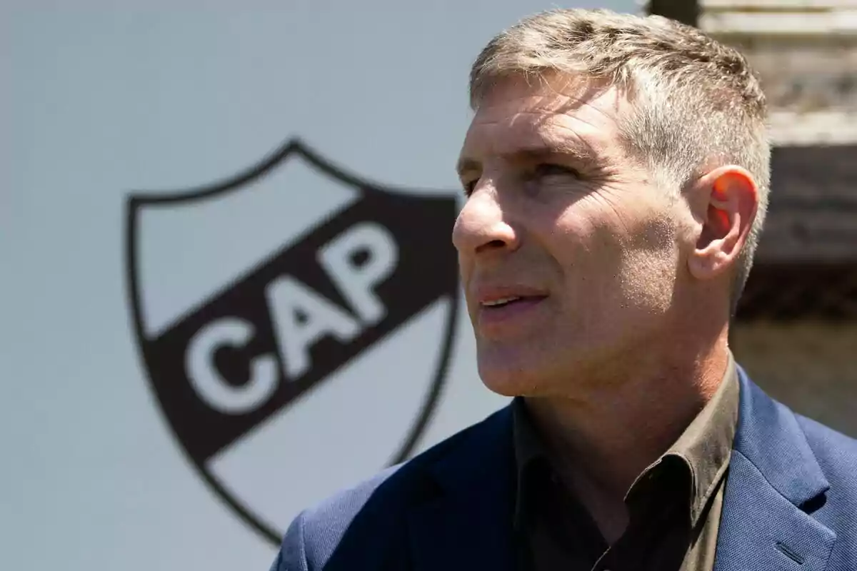 Hombre de cabello corto y canoso con saco azul y camisa oscura, de perfil, frente a un escudo con las letras CAP. Hombre de cabello corto y canoso con saco azul y camisa oscura, de perfil, frente a un escudo con las letras CAP.