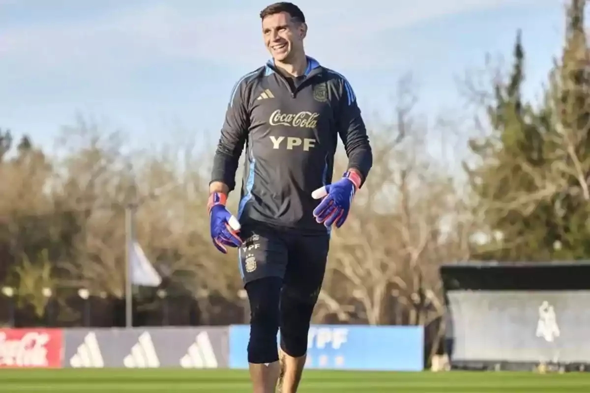 Portero de fútbol sonriente con uniforme de entrenamiento de la selección argentina y guantes azules caminando sobre el césped de una cancha Portero de fútbol sonriente con uniforme de entrenamiento de la selección argentina y guantes azules caminando sobre el césped de una cancha