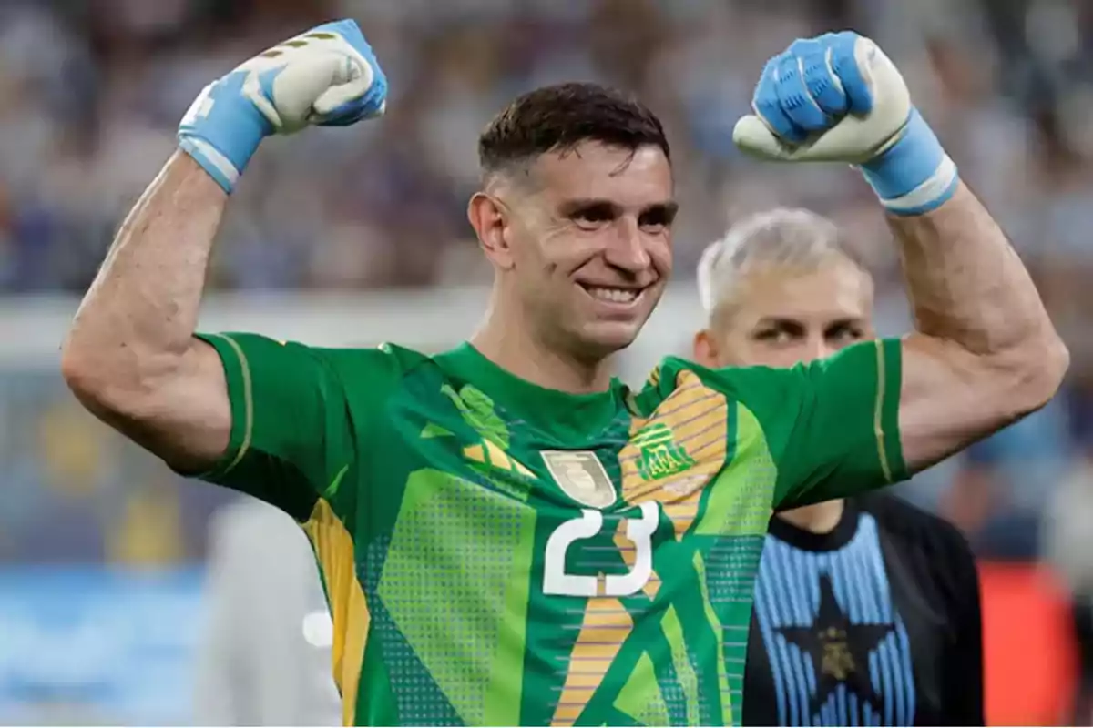 Un futbolista con uniforme verde levanta los brazos en señal de victoria mientras sonríe.