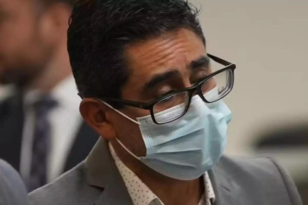 Hombre con gafas y cubrebocas azul claro viste saco gris y camisa blanca con puntos pequeños Hombre con gafas y cubrebocas azul claro viste saco gris y camisa blanca con puntos pequeños