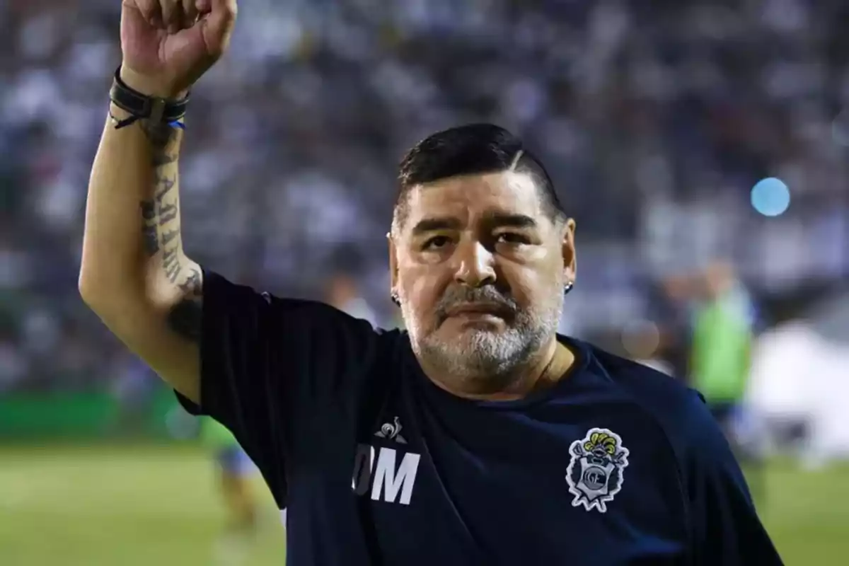 Hombre con barba y cabello corto levantando el brazo en un estadio de fútbol, lleva una camiseta azul oscuro con un escudo y las iniciales DM