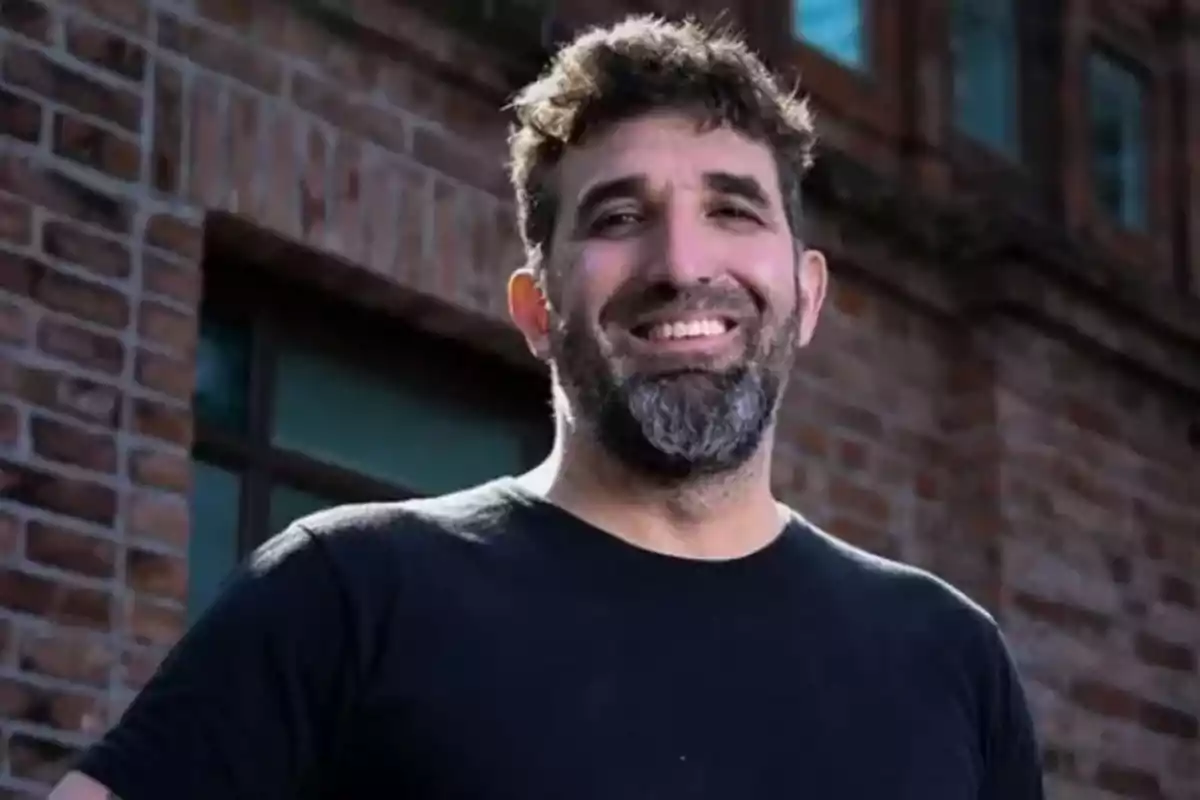 Curly-haired man with a beard wearing a black t-jersey smiling in front of a brick wall Curly-haired man with a beard wearing a black t-jersey smiling in front of a brick wall