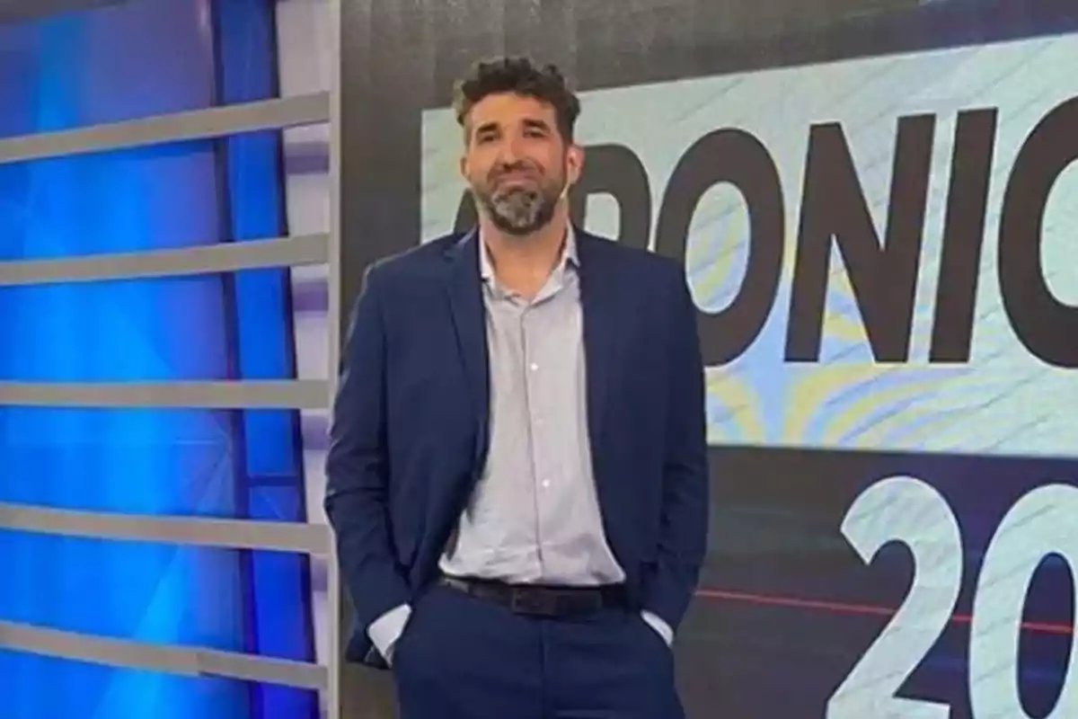 Un hombre con traje azul y camisa clara está de pie frente a un fondo con texto en un estudio de televisión. Un hombre con traje azul y camisa clara está de pie frente a un fondo con texto en un estudio de televisión.