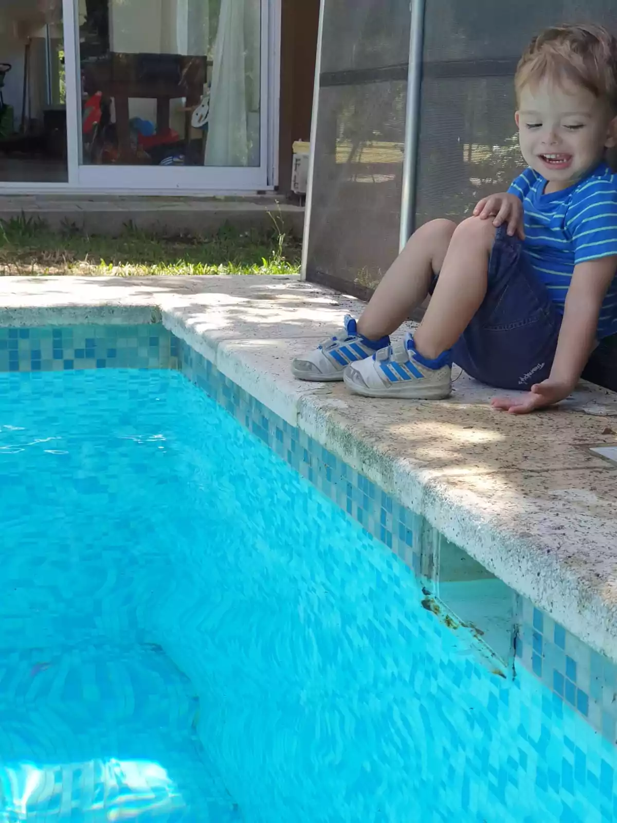 Un niño pequeño con camiseta azul y pantalones cortos está sentado en el borde de una piscina, sonriendo mientras mira el agua. Un niño pequeño con camiseta azul y pantalones cortos está sentado en el borde de una piscina, sonriendo mientras mira el agua.