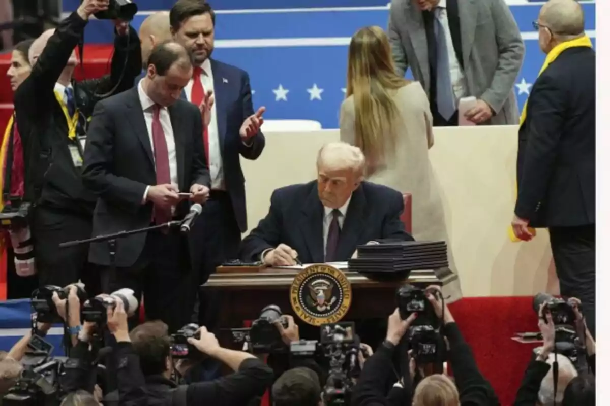 Un hombre mayor con cabello blanco está sentado en un escritorio firmando documentos rodeado de personas y fotógrafos en un evento formal. Un hombre mayor con cabello blanco está sentado en un escritorio firmando documentos rodeado de personas y fotógrafos en un evento formal.