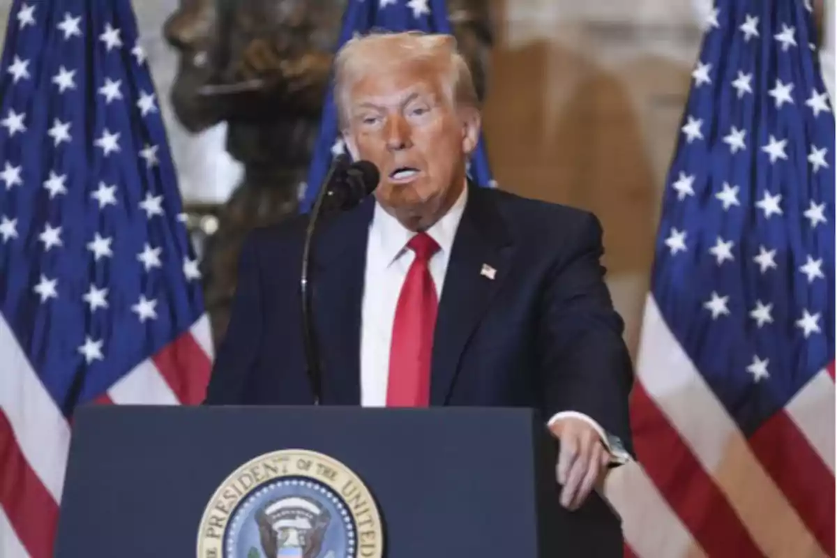 Un hombre de traje oscuro y corbata roja habla frente a un podio con el sello presidencial de los Estados Unidos, con dos banderas estadounidenses de fondo.