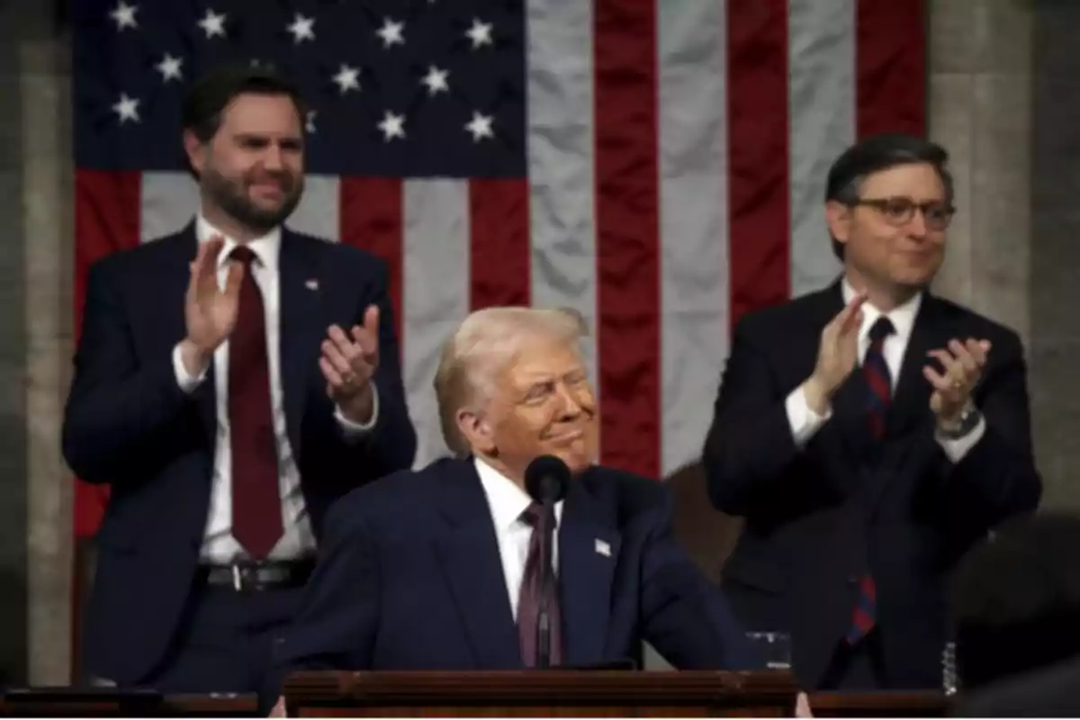 A man on a podium smiles while two people behind him applaud, with a United States flag in the background.