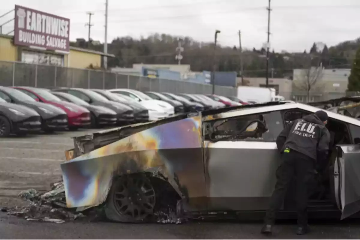 A burned vehicle with a fire department agent inspecting it, parked near a row of cars in an industrial area.