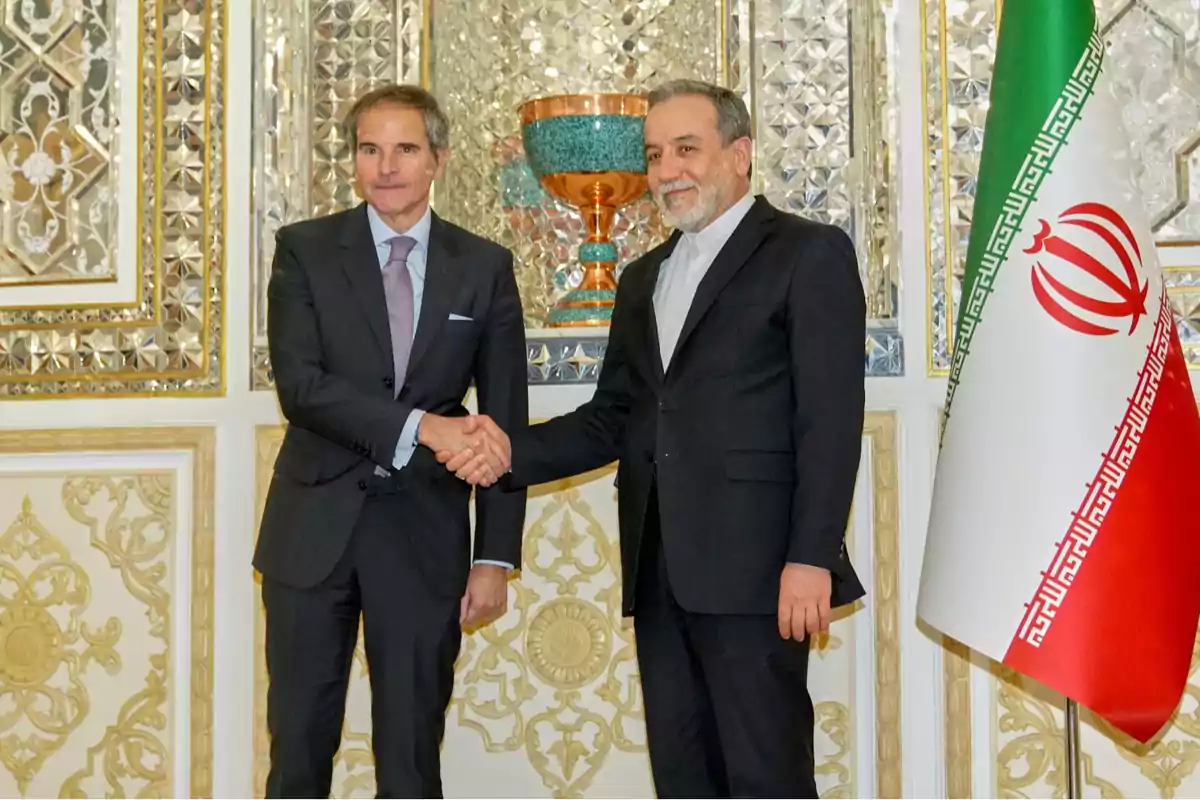 Dos hombres de traje se dan la mano frente a una decoración elaborada y una bandera iraní. Dos hombres de traje se dan la mano frente a una decoración elaborada y una bandera iraní.