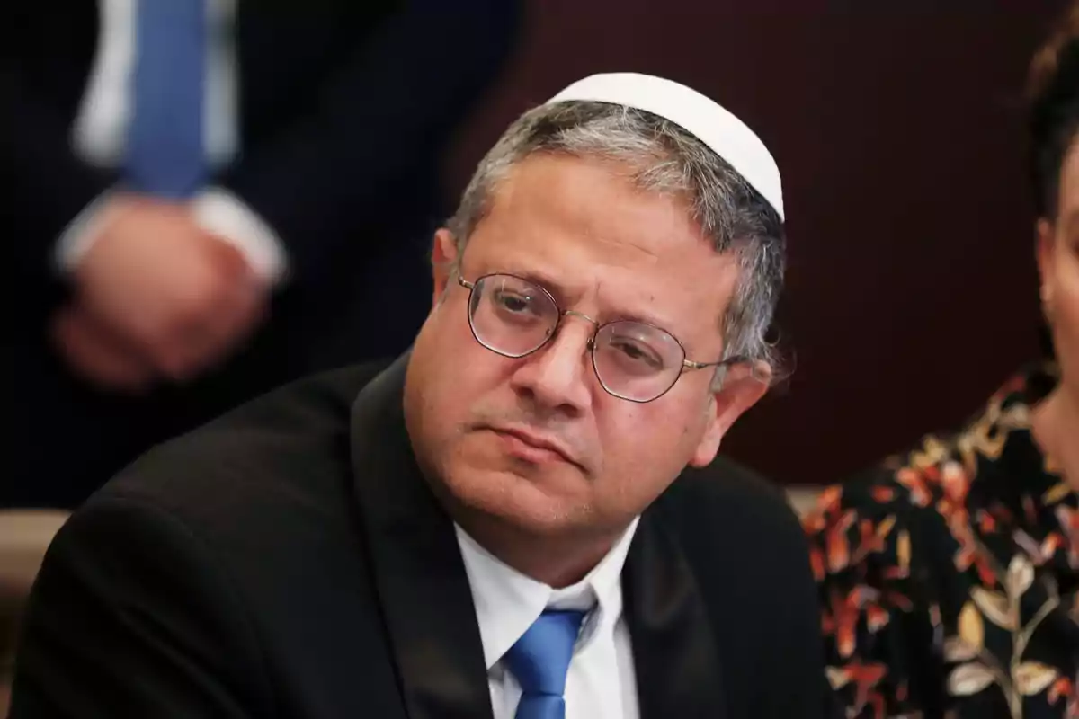Hombre con kipá blanca y gafas sentado en una reunión formal con expresión seria Hombre con kipá blanca y gafas sentado en una reunión formal con expresión seria