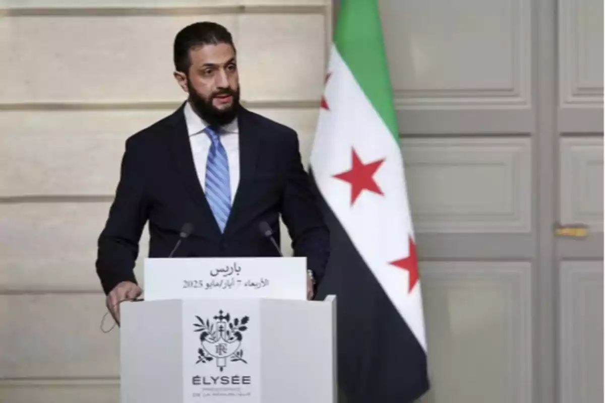 Hombre con barba y traje oscuro hablando en un podio con la bandera de Siria de fondo