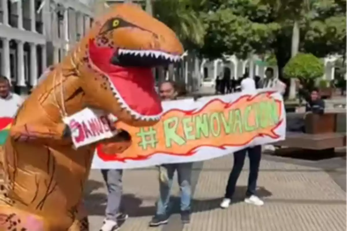 Una persona disfrazada de dinosaurio sostiene un cartel mientras otras personas detrás sostienen una pancarta con el texto 