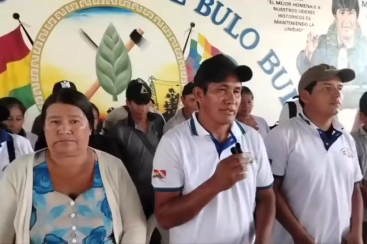 Un grupo de personas vestidas con camisetas blancas se encuentra reunido frente a un mural colorido que incluye una hoja verde y herramientas cruzadas, mientras una persona sostiene un micrófono. Un grupo de personas vestidas con camisetas blancas se encuentra reunido frente a un mural colorido que incluye una hoja verde y herramientas cruzadas, mientras una persona sostiene un micrófono.