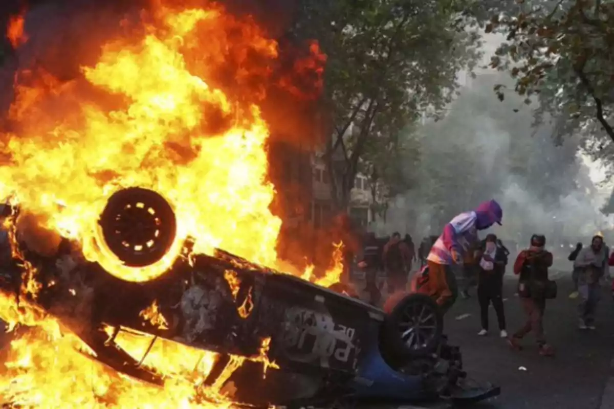 An overturned car in flames on a street with people around, some taking photos and others watching.