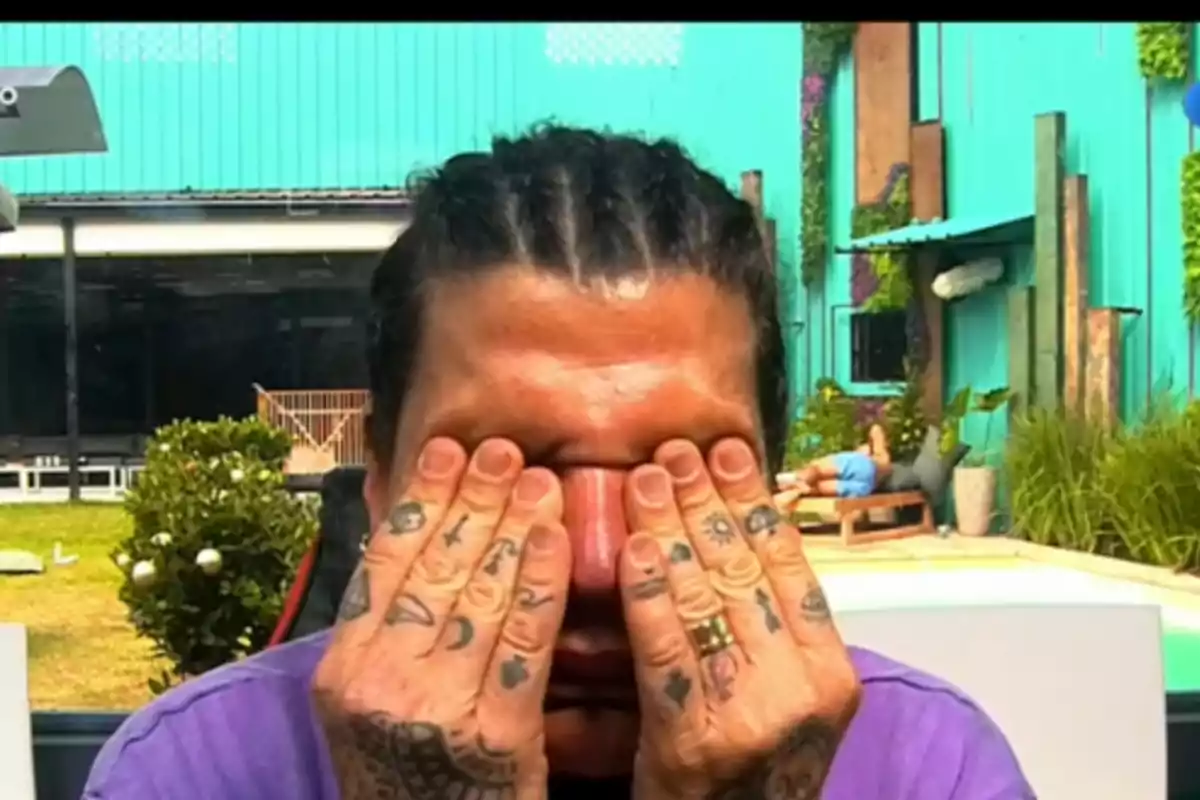 Una persona con trenzas se cubre los ojos con las manos tatuadas en un entorno al aire libre con una piscina y vegetación.