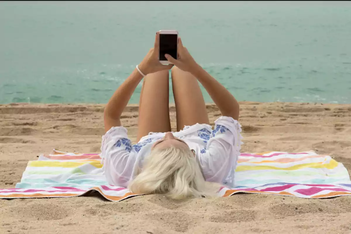 Persona recostada en la playa usando un teléfono móvil sobre una toalla de rayas coloridas frente al mar.