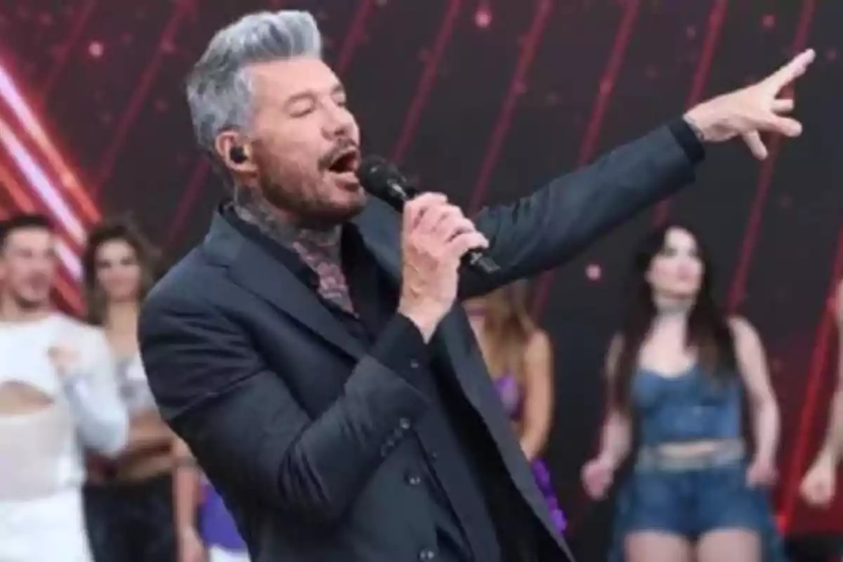 Un hombre con cabello canoso y barba canta en un escenario con un micrófono mientras señala hacia adelante, con personas desenfocadas en el fondo.