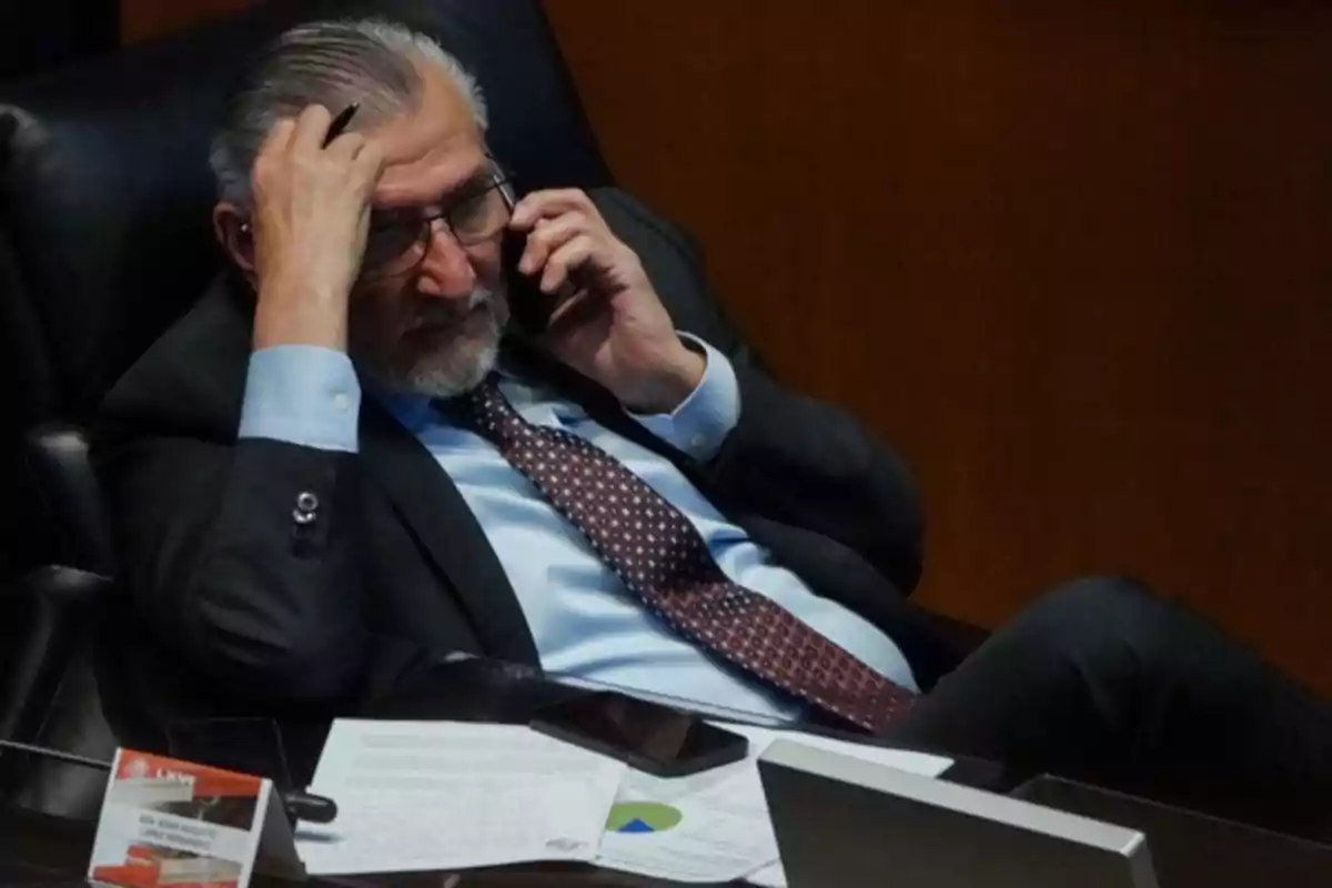 Hombre de traje y corbata sentado en una silla, hablando por teléfono y sosteniéndose la cabeza con una mano, rodeado de documentos y un teléfono móvil sobre la mesa Hombre de traje y corbata sentado en una silla, hablando por teléfono y sosteniéndose la cabeza con una mano, rodeado de documentos y un teléfono móvil sobre la mesa