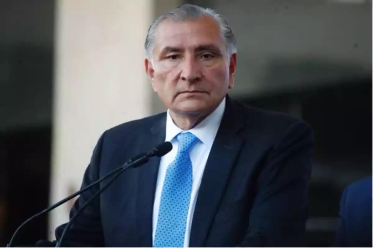 Hombre de cabello canoso con traje oscuro y corbata azul hablando frente a un micrófono