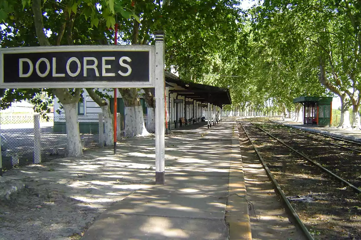Andén de la estación de tren Dolores rodeado de árboles y vías vacías en un día soleado
