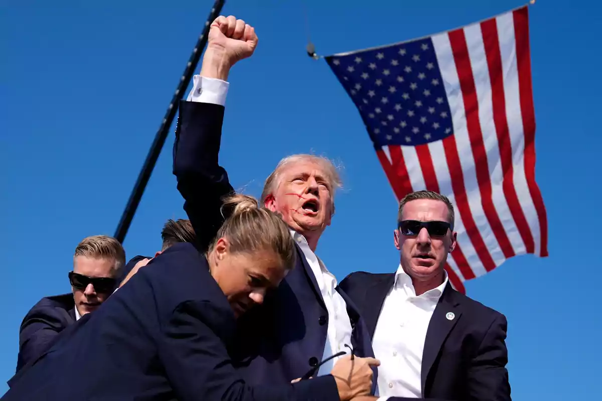 A group of people surrounds a man who raises his fist, with a United States flag waving in the background.