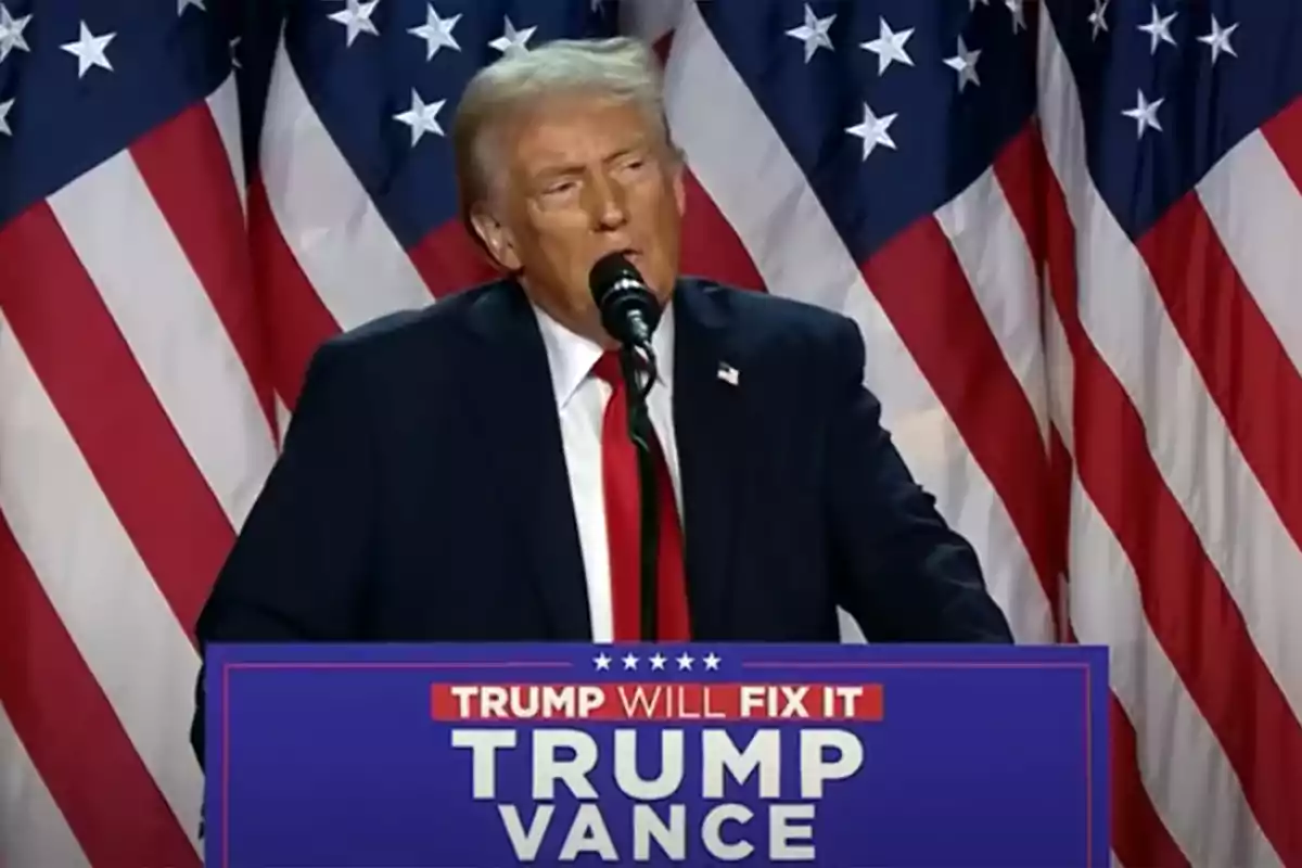 Hombre de cabello rubio con traje oscuro y corbata roja hablando en un podio azul con banderas de Estados Unidos de fondo Hombre de cabello rubio con traje oscuro y corbata roja hablando en un podio azul con banderas de Estados Unidos de fondo
