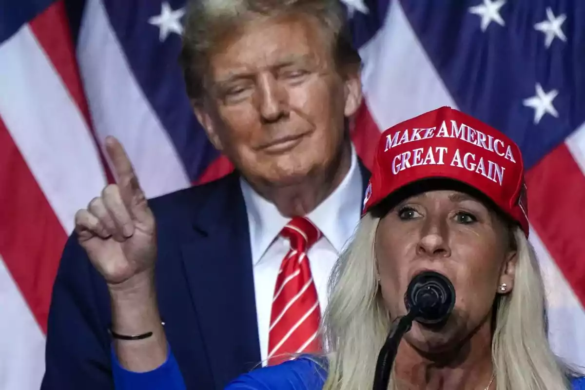 A person with a red cap speaking into a microphone with a male figure in the background and United States flags behind. A person with a red cap speaking into a microphone with a male figure in the background and United States flags behind.