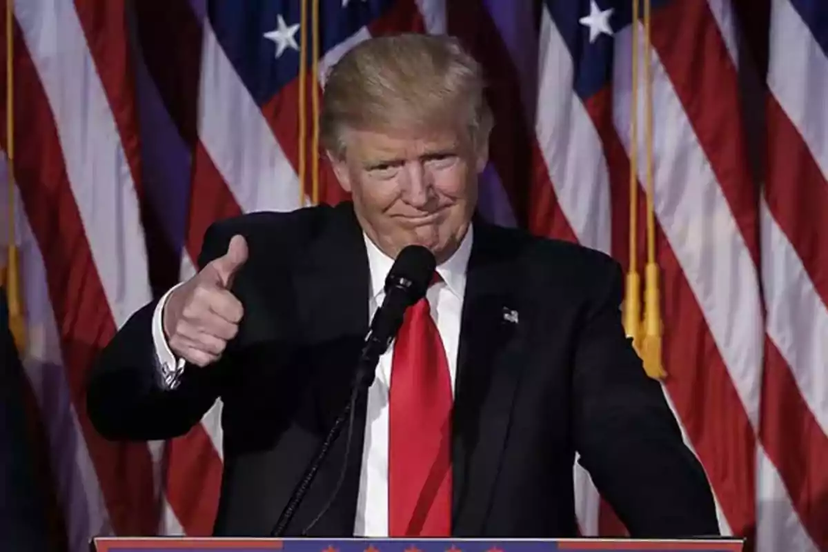 A man in a dark suit with a red tie gives a thumbs-up gesture in front of a background of flags.