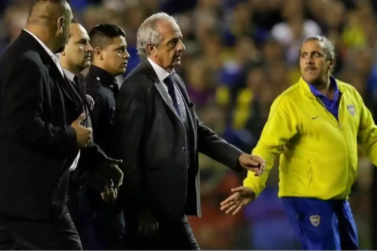 Un grupo de hombres camina en un estadio, uno de ellos lleva un traje y otro un uniforme deportivo amarillo.