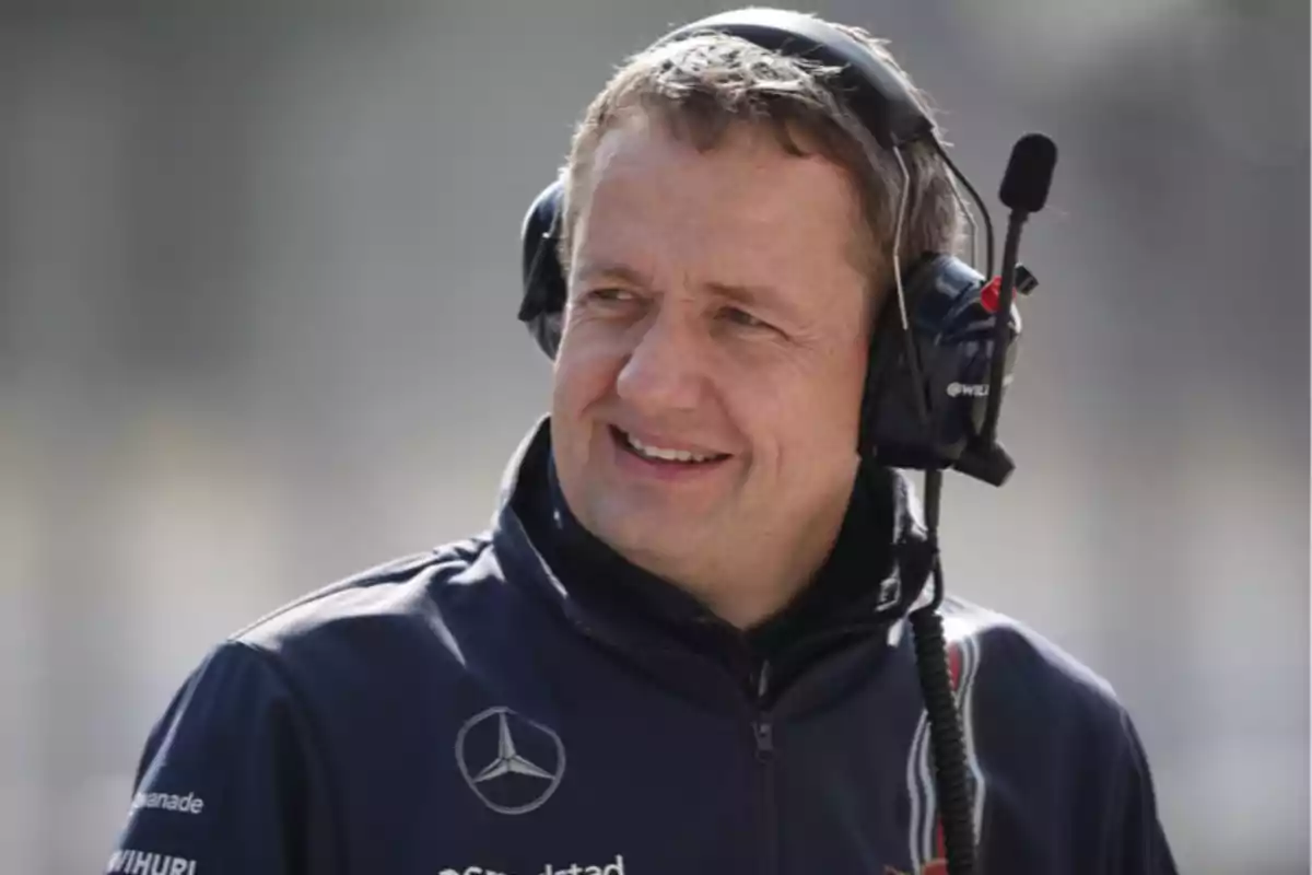 Man with headphones and microphone wearing a blue jacket with a Mercedes logo smiles outdoors