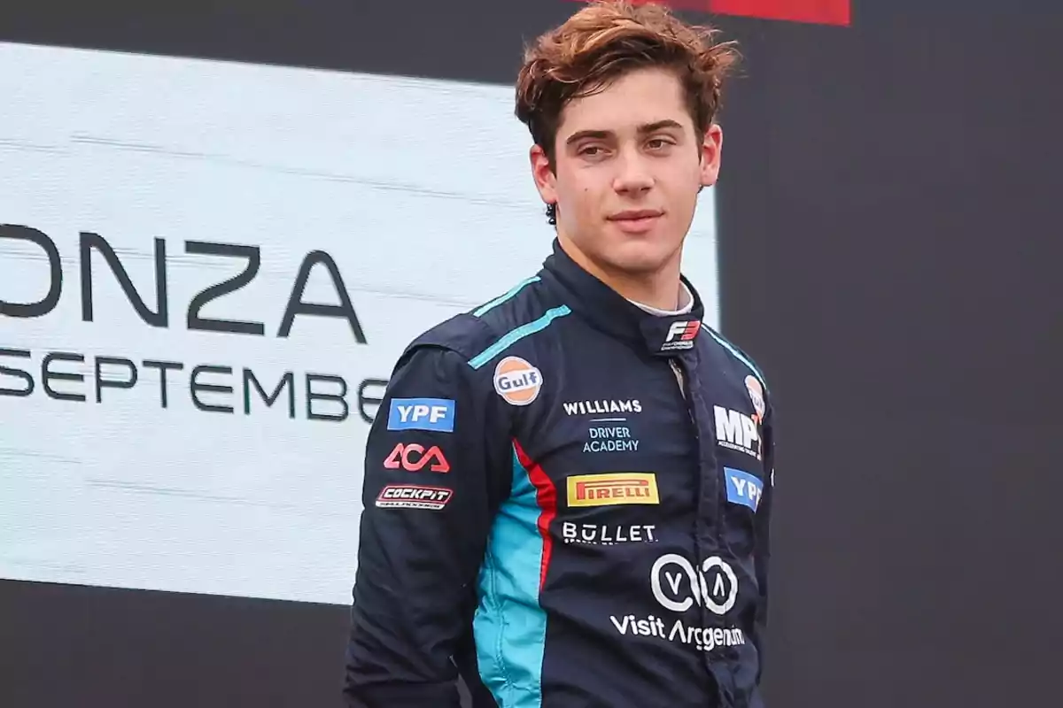 Un piloto de carreras con un traje azul oscuro lleno de logotipos de patrocinadores, de pie frente a un cartel que dice "Monza September".