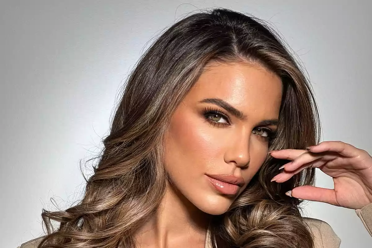 Long-haired, wavy-haired woman with flawless makeup looking at the camera and touching her face with one hand
