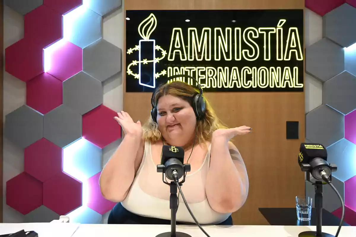 Una persona con audífonos está sentada frente a un micrófono en un estudio de grabación con el logo de Amnistía Internacional en el fondo.