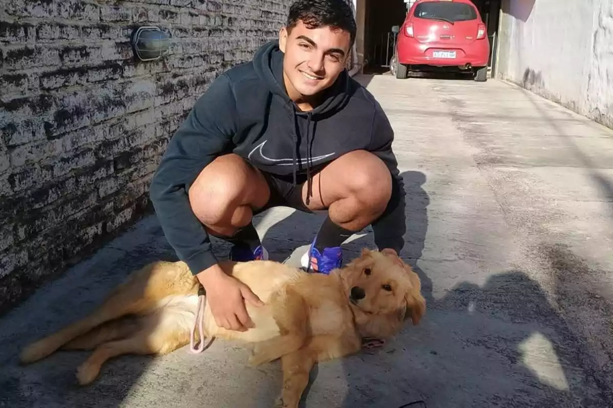 Smiling young man crouching down, petting a golden dog lying on the ground on a street next to a brick wall, with a red car in the background Smiling young man crouching down, petting a golden dog lying on the ground on a street next to a brick wall, with a red car in the background